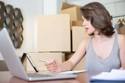 An online store owner working through orders on a laptop, shipping boxes stacked behind her